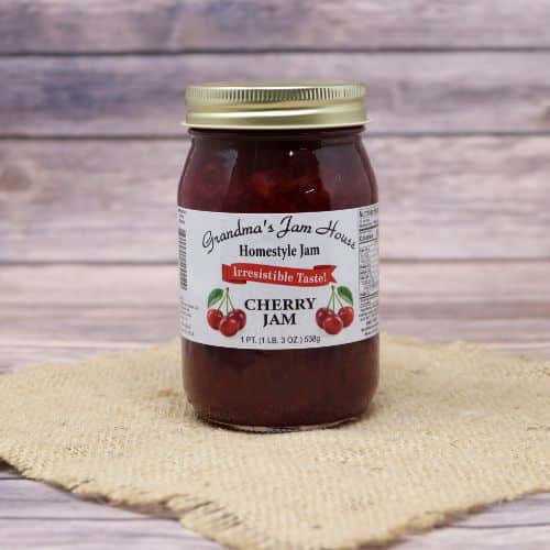A pint size glass jar of Grandma's Jam House Cherry Jam, on rustic burlap with a wooden background.