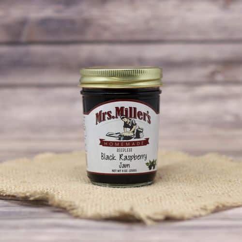 A glass jar of Mrs. Miller's Seedless Black Raspberry Jam, on rustic burlap with a wooden background.