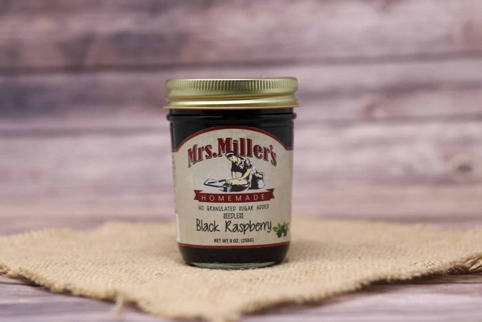 A 9 oz glass jar of Mrs. Miller's Seedless Black Raspberry Jam No Sugar Added, on rustic burlap with a wooden background.