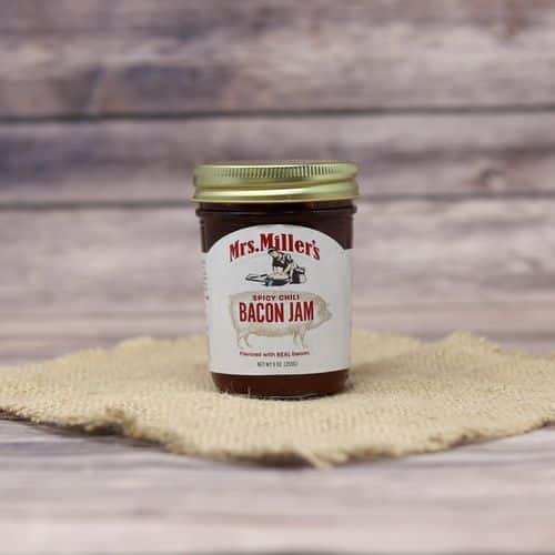 A glass jar of Mrs. Miller's Spicy Chili Bacon Jam, on rustic burlap with a wooden background.