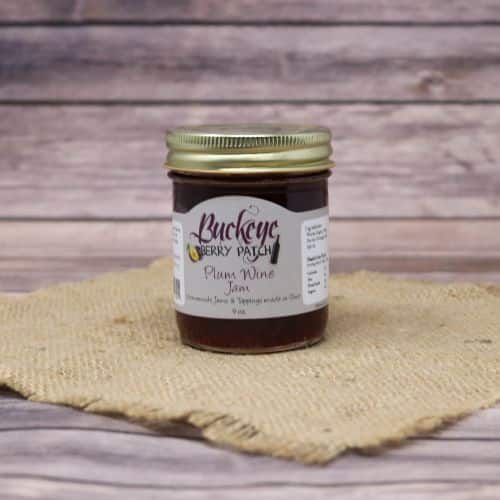 A glass jar of Buckeye Berry Patch Plum Wine Jam, on rustic burlap with a wooden background.