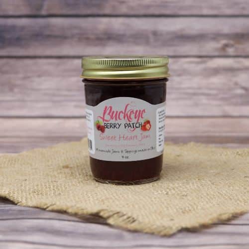 A glass jar of Buckeye Berry Patch Sweet Heart Jam, on rustic burlap with a wooden background.