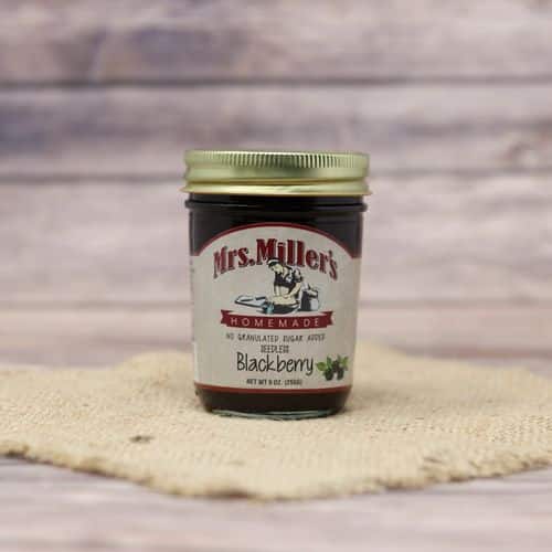 A 9 oz glass jar of Mrs. Miller's No Sugar Added Seedless Blackberry Jam, on rustic burlap with a wooden background.
