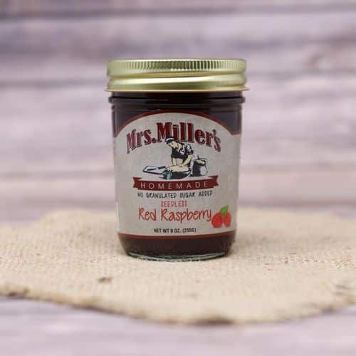 A 9 oz glass jar of Mrs. Miller's No Sugar Added Seedless Red Raspberry Jam, on rustic burlap with a wooden background.