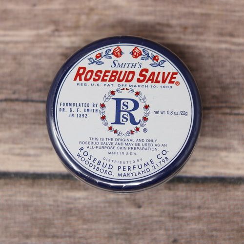 A 0.8 oz metal container of Rosebud Salve, set against a rustic wood background.