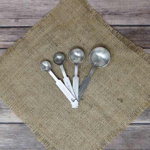 A set of 4 stainless steel measuring spoons, laying on rustic burlap with a wooden background.
