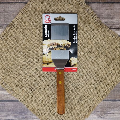 A Chef Craft spatula with a wooden handle and stainless steel blade, laying on rustic burlap with a wooden background.
