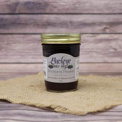 A 9oz glass jar of Buckeye Berry Patch Blackberry Preserves, on rustic burlap with a wooden background.