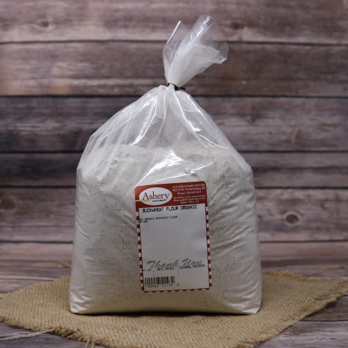 Bag of Ashery organic buckwheat flour, sealed and standing on a burlap mat with a weathered wood background.
