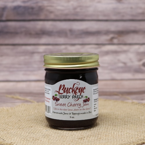 A glass jar of Buckeye Berry Patch Sweet Cherry Jam, on rustic burlap with a wooden background.