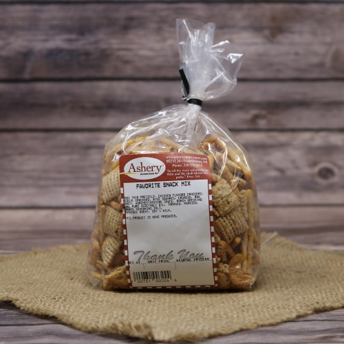 A sealed bag of Ashery's Favorite Snack Mix on a straw mat with a wooden background, displaying an assortment of snacks within