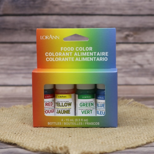 A pack of LorAnn liquid food coloring in four colors—red, yellow, green, and blue—on a straw mat with a wooden texture in the background.