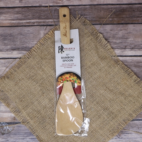 A Helen's Asian Kitchen bamboo spoon, wrapped in plastic packaging, lies on rustic burlap with a wooden backdrop.