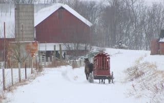 A snowy winter scene with a red barn in the background and a horse and wagon in the foreground.