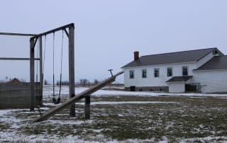 A winter scene of a school yard with a swing set and seesaw in the foreground and a schoolhouse in the background.