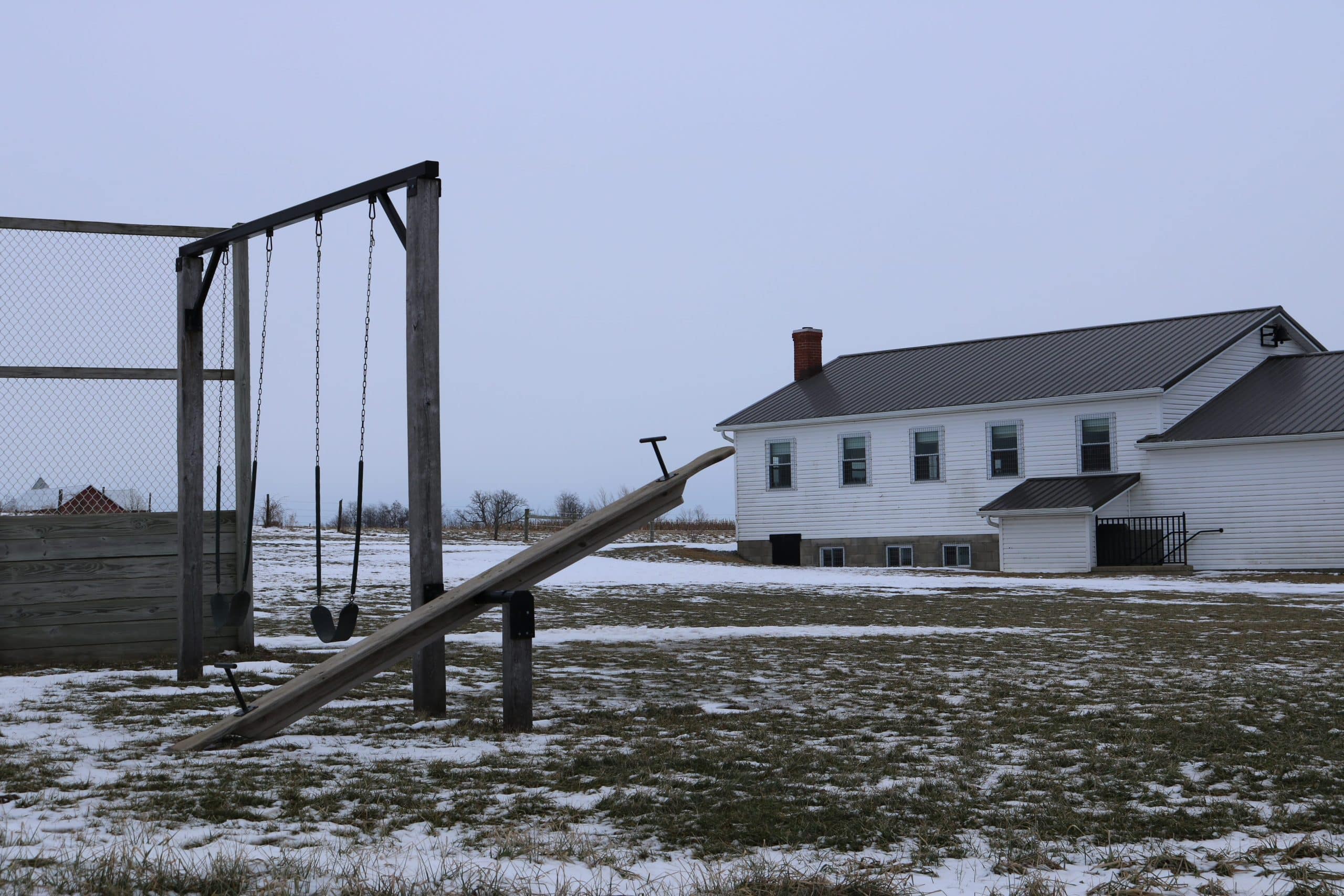 A winter scene of a school yard with a swing set and seesaw in the foreground and a schoolhouse in the background.