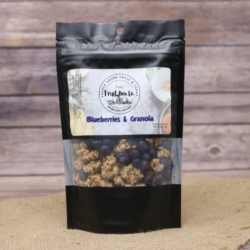 A black resealable bag with a clear window revealing Blueberries And Granola Freeze Dried on a rustic burlap with a wooden background.