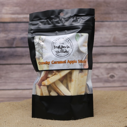 A black resealable bag with a clear window revealing Crunchy Caramel Apple Sticks Freeze Dried on a rustic burlap with a wooden background.