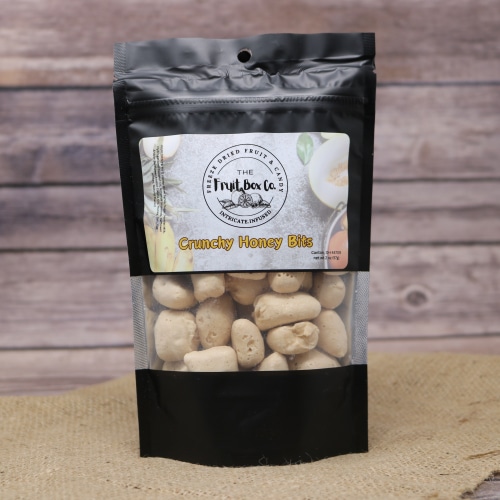 A black resealable bag with a clear window revealing Crunchy Honey Bits Freeze Dried on a rustic burlap with a wooden background.
