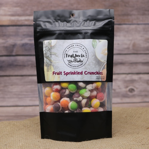 A black bag with a clear window revealing Fruit Sprinkled Crunchies Freeze Dried on a rustic burlap with a wooden background.