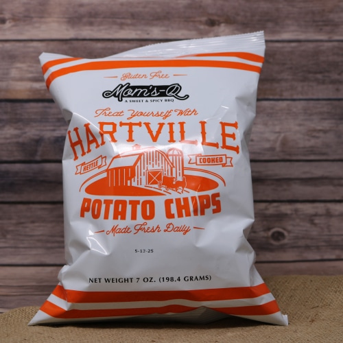 A white bag with orange writing of Hartville Potato Chips BBQ on rustic burlap with a wooden background.