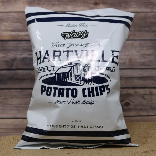 A white bag with blue writing of gluten free Hartville Potato Chips Wavy on rustic burlap with a wooden background.