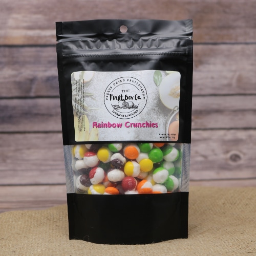 A black bag with a clear window revealing Rainbow Crunchies Freeze Dried on a rustic burlap with a wooden background.