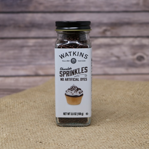 A small glass shaker container with a removable lid of Watkins Chocolate Sprinkles, these sprinkles have no artificial dyes, on rustic burlap with a wooden background.