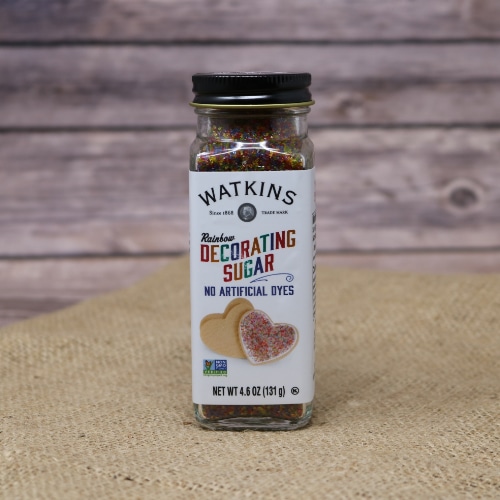 A small glass shaker container with a removable lid of Watkins Rainbow Decorating Sugar, these sprinkles have no artificial dyes, on rustic burlap with a wooden background.