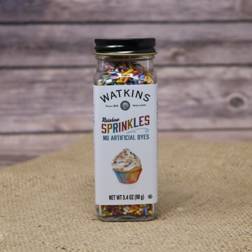 A small glass shaker container with a removable lid of Watkins Rainbow Sprinkles, these sprinkles have no artificial dyes, on rustic burlap with a wooden background.