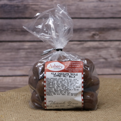 A clear plastic bag of Milk Chocolate Triple Dipped Malt Balls sealed with a black twist tie on rustic burlap with a wooden background.