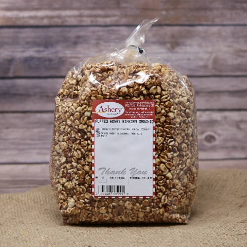 A clear bag of Puffed Honey Einkorn Organic sealed with a twist tie on rustic burlap with a wooden background.