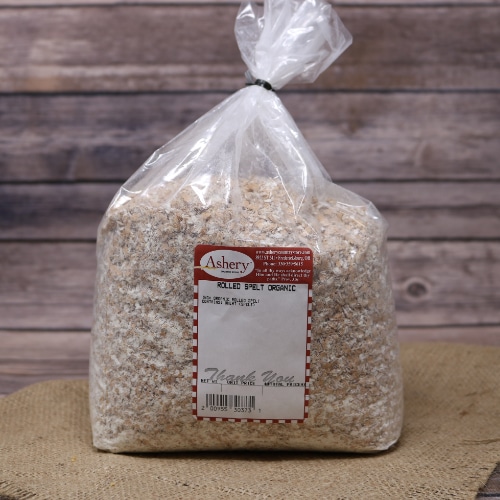 A clear bag of Rolled Spelt Organic sealed with a twist tie on rustic burlap with a wooden background.