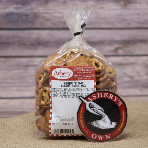 A clear plastic bag of Sesame Bagel Snack Mix Ashery's Own sealed with a black twist tie on rustic burlap with a wooden background.