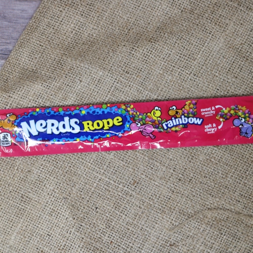 A long thin pink package containing a Nerd Rope Rainbow on rustic burlap with a wooden background.