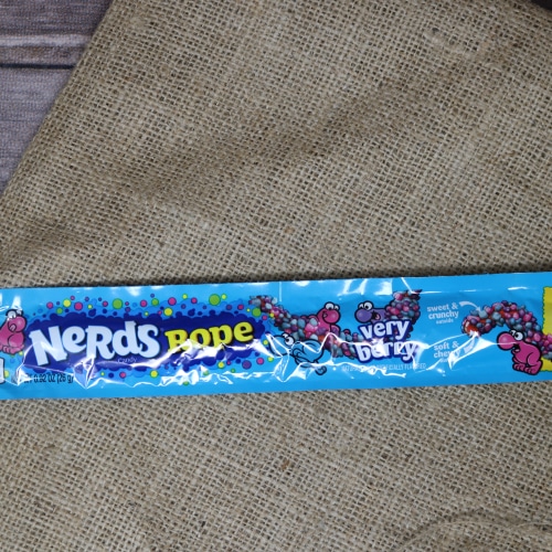 A long thin blue package containing a Nerd Rope Very Berry on rustic burlap with a wooden background.
