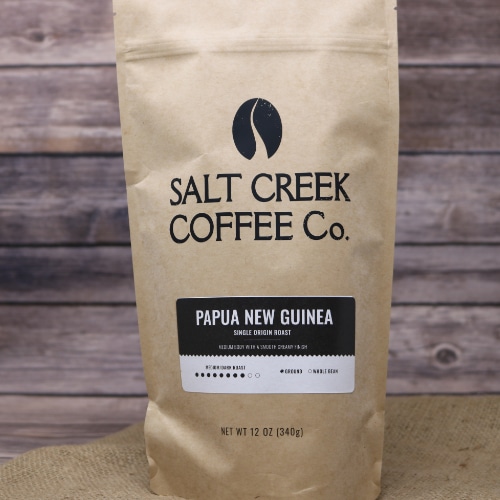 A tall brown bag with black lettering of Papua New Guinea Ground Coffee 12 oz on rustic burlap with a wooden background.