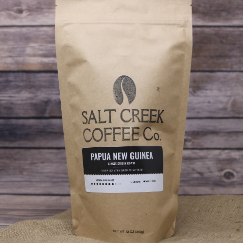 A tall brown bag with black lettering of Papua New Guinea Whole Bean Coffee 12 oz on rustic burlap with a wooden background.