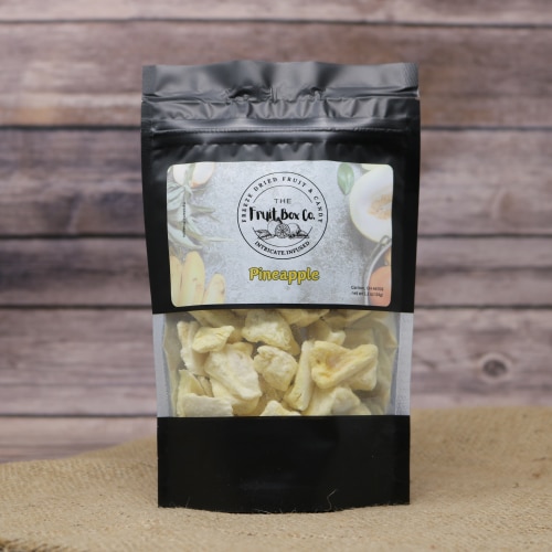 A black bag with a clear window revealing Freeze Dried Pineapple on a rustic burlap with a wooden background.